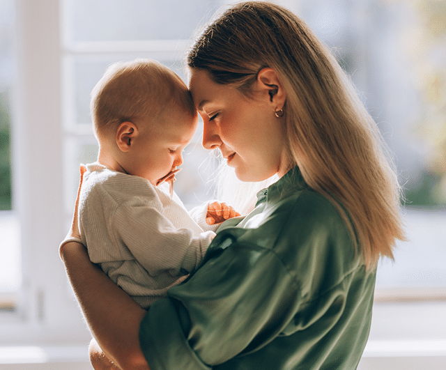 A young mother in a green shirt tenderly holds her baby, touching foreheads in a warm, sunlit room.