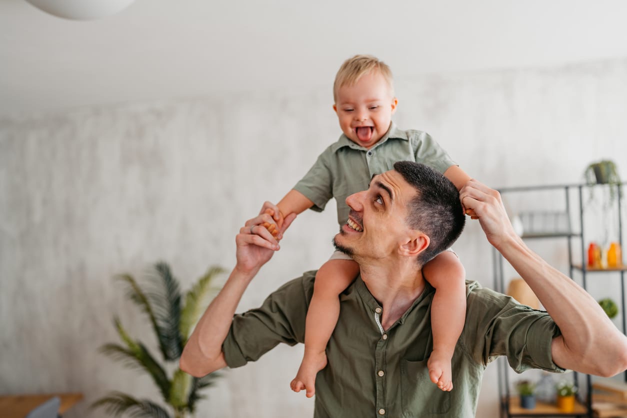 Young Father Carrying His Child With Down Syndrome On The Shoulders In The Living Room At Home