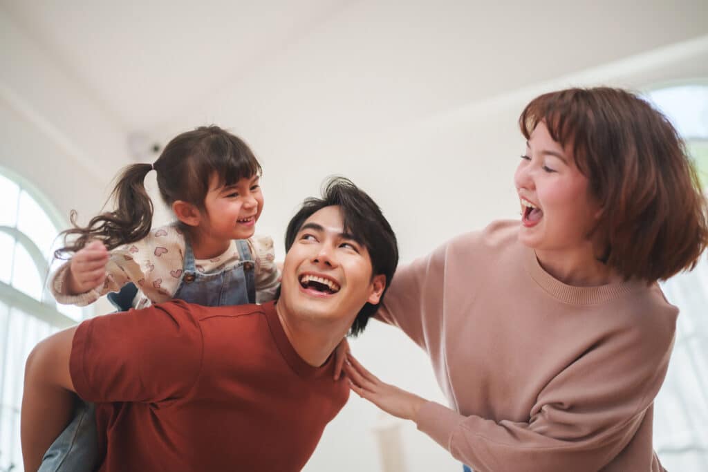Asian daughter riding on parents back while laughing together at home