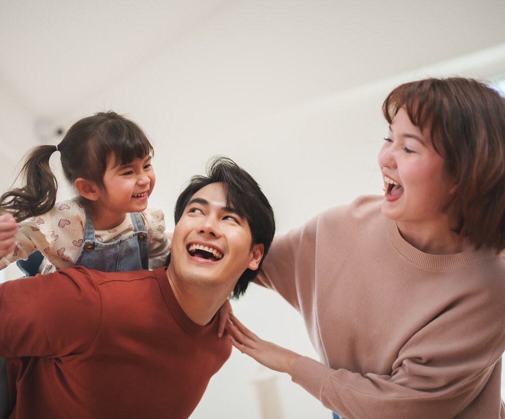 A joyful Asian family laughing together, with a father giving his young daughter a piggyback ride while the mother smiles ...